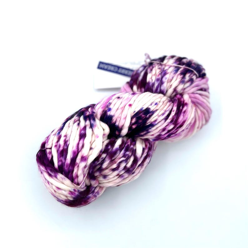 May include: A skein of thick, hand-dyed yarn featuring a variegated pattern of deep purple, magenta, and cream white. The yarn has a soft, twisted texture and includes a small white paper tag with dark blue text attached to the top section.
