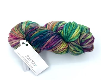 May include: A skein of super bulky Rasta yarn featuring a variegated color palette of deep purple, emerald green, and golden yellow. A white rectangular product tag is attached, displaying the text RASTA, 90 yd, and 100% Merino Wool.