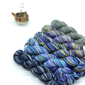May include: Four skeins of yarn in shades of blue, black, and white with a striped pattern. The yarn is arranged in a fan shape.