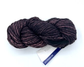 May include: A skein of thick, dark purple and black variegated yarn with a twisted texture. A white paper label is attached to the yarn, featuring the text RASTA and 100 SINGLE LOT printed in dark blue ink on a blue background.