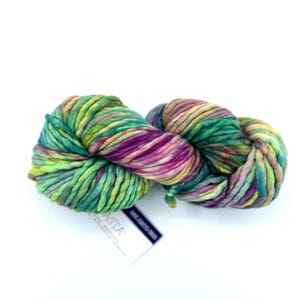 May include: A skein of thick, chunky yarn featuring a variegated color palette of vibrant green, deep purple, and soft yellow tones. The yarn is twisted into a figure-eight shape and includes a small white paper tag labeled ASTA.