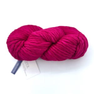 May include: A skein of chunky, vibrant magenta yarn with a thick, twisted texture. The yarn is wound into a large, soft oval shape. A small white paper tag with partially visible black text is attached to the side of the yarn bundle.