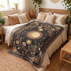 Könnte beinhalten: Eine schwarze Webdecke mit einem zentralen Sonnen- und Mondmotiv, umgeben von floralen und botanischen Mustern in Gold-, Creme- und Brauntönen. Die rechteckige Decke ist mit einem Fransenrand versehen und detailliert gestaltet.