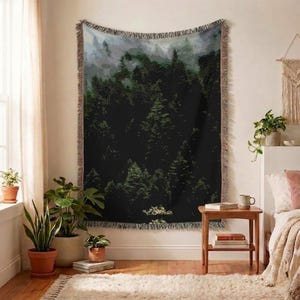 May include: A rectangular woven throw blanket featuring a dark forest landscape with dense green pine trees and misty gray mountains in the background. The blanket has a decorative fringed edge on all four sides and a textured weave.