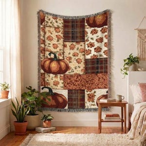 Op de afbeelding: Een rechthoekige geweven plaid met een patchworkontwerp van pompoenen, herfstbladeren en ruitpatronen in tinten oranje, bruin en crème. De deken heeft een gefranjerde rand en hangt verticaal aan een muur.