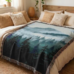 Puede incluir: Una manta tejida que presenta una ilustración de paisaje de un bosque denso con pinos altos en tonos verde oscuro, azul y negro, que se transforma en una cordillera brumosa de color azul claro bajo un cielo pálido, con bordes de flecos.