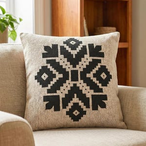 May include: A beige decorative pillow with a large black geometric snowflake design. The pillow is on a beige armchair, with a window and a plant in the background. The pillow is a square shape.