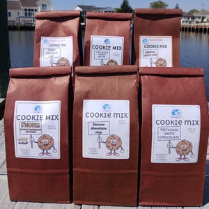 May include: Six brown paper bags of cookie mix arranged in two rows. Each bag features a white label with the text "COOKIE MIX" and a cartoon cookie character holding a sign with flavor names: Cherry Chocolate Chip, Chocolate Peanut Butter, Pumpkin White Chip, S'mores, Banana Chocolate Chip, and Pistachio White Chocolate.