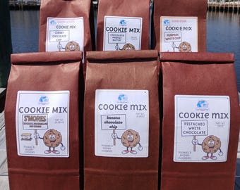 Könnte beinhalten: Sechs braune Papiertüten mit Keks-Mix werden im Freien präsentiert. Jede Tüte hat ein weißes Etikett mit den Worten "Cookie Mix" und einer Cartoon-Keksfigur. Zu den Geschmacksrichtungen gehören Cherry Chocolate Chip und S'mores.