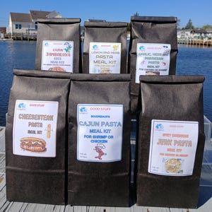 May include: A collection of six dark brown paper bags, each labeled with a different pasta meal kit. The kits include Cajun Jambalaya, Lemon and Herb Pasta, Garlic Herb and Parmesan Pasta, Cheesesteak Pasta, Cajun Pasta, and Spicy Caribbean Jerk Pasta.
