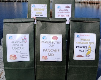 Könnte beinhalten: Fünf dunkelgrüne Papiertüten mit Pfannkuchenmischung sind zu sehen. Die Etiketten tragen Aufschriften wie "Birthday Cake Pancake", "Chocolate Chip Pancake", "Grandma's Apple Pie Pancake Mix", "Peanut Butter Cup Pancake Mix" und "Banana Chocolate Chip Pancake Mix".