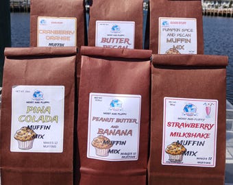 Könnte beinhalten: Sechs braune Papiertüten mit Muffin-Mix werden im Freien gezeigt. Die Geschmacksrichtungen sind Cranberry Orange, Butter Pecan, Pumpkin Spice and Pecan, Pina Colada, Peanut Butter and Banana und Strawberry Milkshake. Jede Tüte hat ein Etikett.