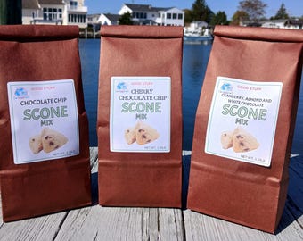 Könnte beinhalten: Drei braune Papiertüten mit Scone-Mischung und weißen Etiketten. Auf den Etiketten steht "Chocolate Chip Scone Mix", "Cherry Chocolate Chip Scone Mix" und "Cranberry, Almond and White Chocolate Scone Mix". Jede Tüte hat ein Nettogewicht von 567 Gramm.