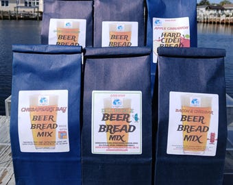 Könnte beinhalten: Eine Auswahl an dunkelblauen Papiertüten mit Bierbrotmischung. Jede Tüte hat ein Etikett mit "Beer Bread" und einer Bierkrug-Grafik. Zu den Geschmacksrichtungen gehören Apfel-Zimt, Dill-Pickle und Bacon & Cheddar. Die Tüten sind ausgestellt.