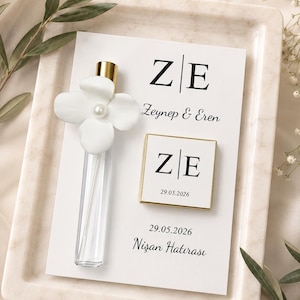 May include: A white card with the initials "ZE" and the names "Zeynep & Eren" and the date "29.05.2026". A small glass vial with a white flower and pearl accent is on the card. The card is on a marble tray.
