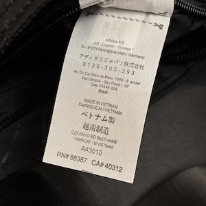 Peut inclure: Gros plan sur une &eacute;tiquette blanche avec du texte noir, attach&eacute;e &agrave; un tissu gris fonc&eacute;. L'&eacute;tiquette comprend le logo Adidas AG, l'adresse et des informations de fabrication. Le texte comprend "MADE IN VIETNAM".