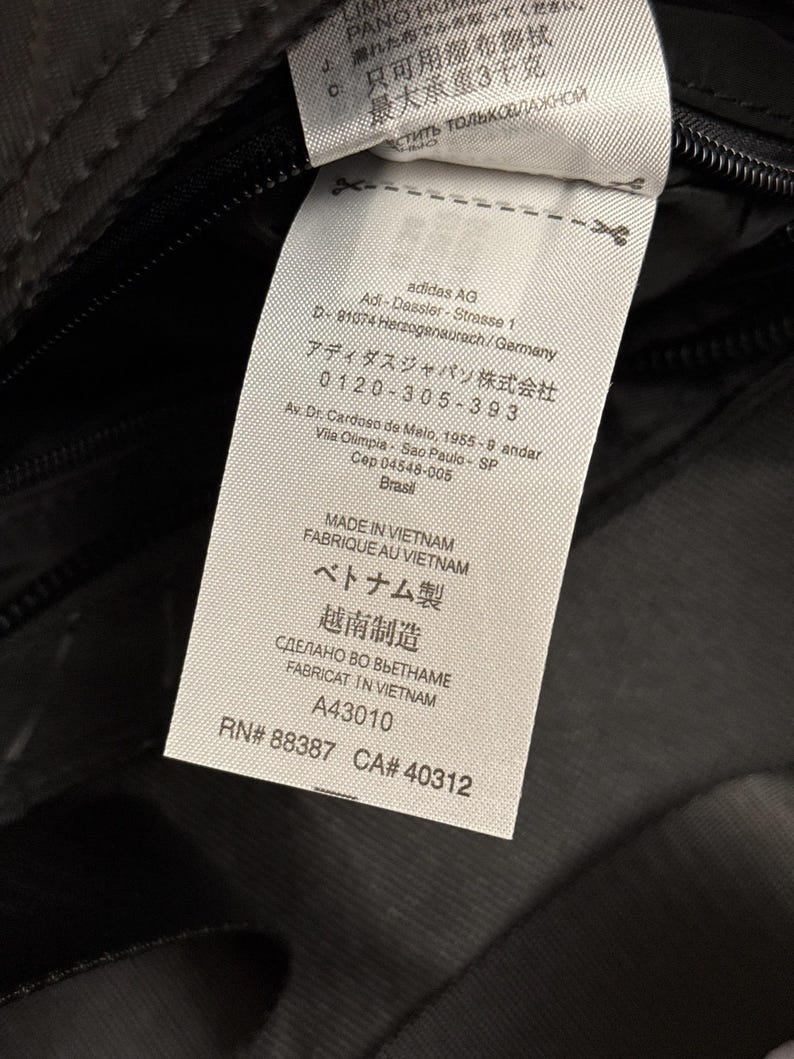 Peut inclure: Gros plan sur une &eacute;tiquette blanche avec du texte noir, attach&eacute;e &agrave; un tissu gris fonc&eacute;. L'&eacute;tiquette comprend le logo Adidas AG, l'adresse et des informations de fabrication. Le texte comprend "MADE IN VIETNAM".