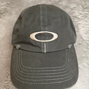 May include: A charcoal grey baseball cap featuring a white embroidered Oakley logo on the front panel. The cap has a curved brim with visible stitching and intentional distressed detailing along the edges of the brim and the front crown area.