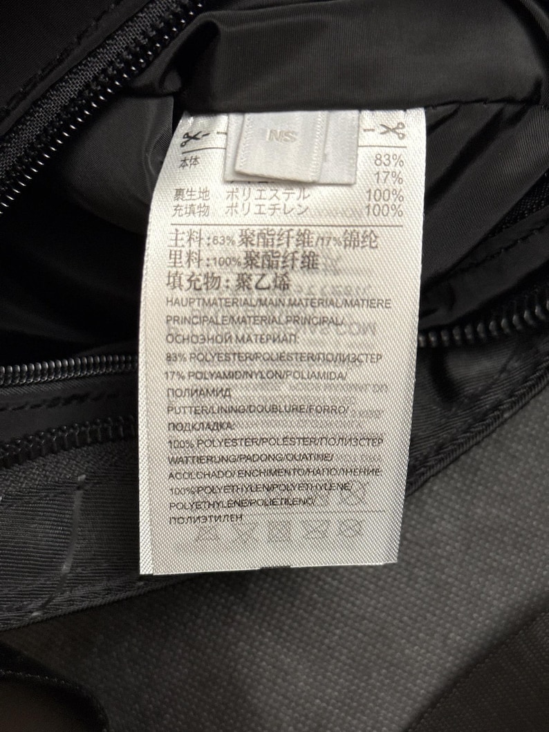 Peut inclure: Gros plan sur un tissu noir avec une &eacute;tiquette blanche attach&eacute;e. L'&eacute;tiquette contient du texte en plusieurs langues, dont le japonais et l'anglais, d&eacute;taillant la composition du tissu, avec des pourcentages de polyester et d'autres mat&eacute;riaux.