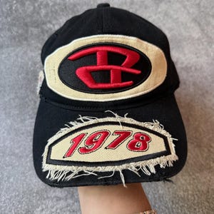 Puede incluir: Gorra de béisbol negra con un logotipo rojo y crema y el año "1978" bordado en la parte delantera. La gorra tiene una visera curva y un diseño clásico de seis paneles.