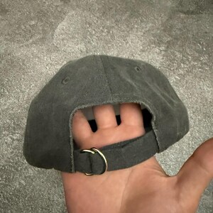 May include: A charcoal gray baseball cap featuring a fabric strap closure at the back. The strap is secured with a gold-toned metal D-ring buckle. The cap has a structured crown with visible stitching and small ventilation eyelets on the top panels.