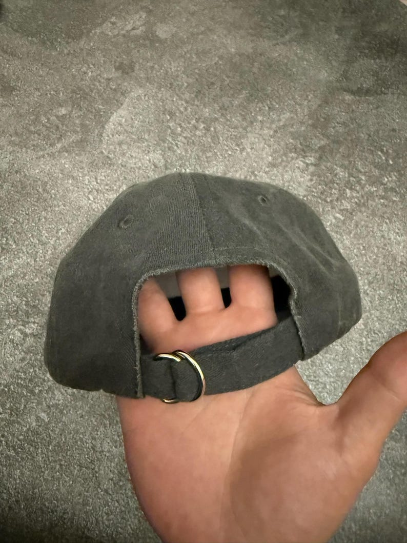 May include: A charcoal gray baseball cap featuring a fabric strap closure at the back. The strap is secured with a gold-toned metal D-ring buckle. The cap has a structured crown with visible stitching and small ventilation eyelets on the top panels.