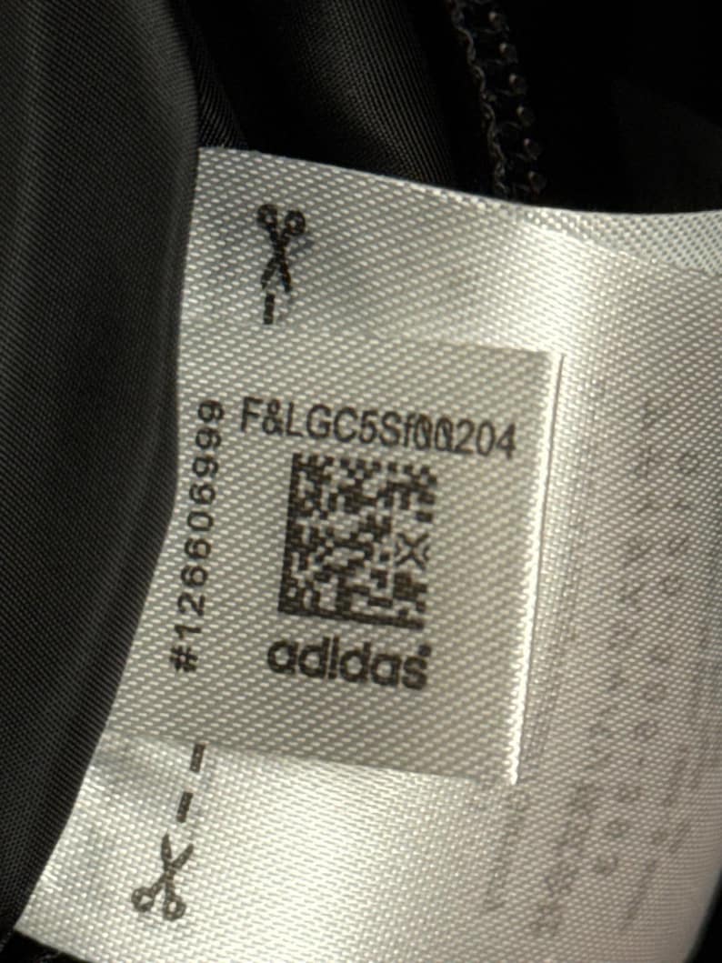 Peut inclure: Gros plan sur une &eacute;tiquette blanche avec le logo Adidas et le texte "F&LGC5Sf00204". L'&eacute;tiquette comprend &eacute;galement un code QR et le num&eacute;ro "126606999". Des ic&ocirc;nes de ciseaux sont imprim&eacute;es sur l'&eacute;tiquette.