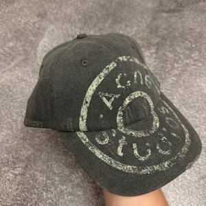 May include: A dark gray baseball cap featuring a distressed, circular light green graphic print that reads Acne Studios. The cap has a structured crown, a curved brim, and stitched eyelets on the top panels, showcasing a worn-in texture.