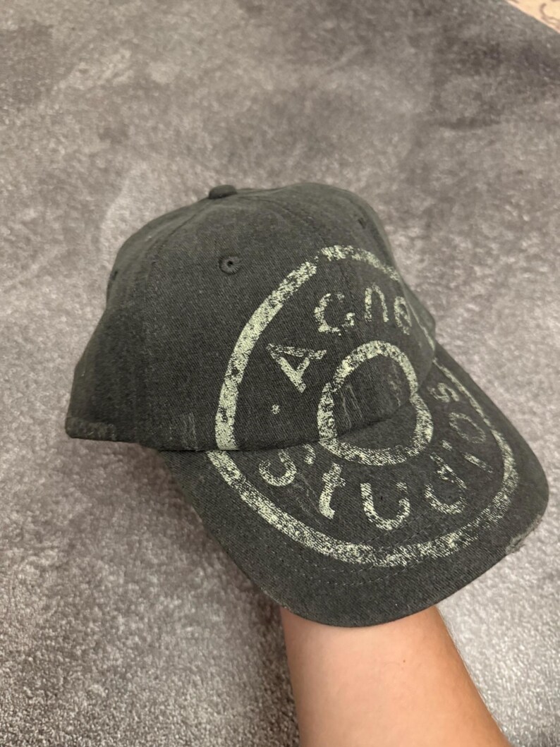 May include: A dark gray baseball cap featuring a distressed, circular light green graphic print that reads Acne Studios. The cap has a structured crown, a curved brim, and stitched eyelets on the top panels, showcasing a worn-in texture.