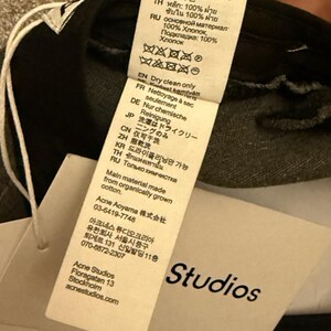 May include: A white fabric care label attached to a black garment, featuring multi-language text including 'Dry clean only' and 'Main material made from organically grown cotton.' A separate white paper tag displays the brand name 'Studios' in black.