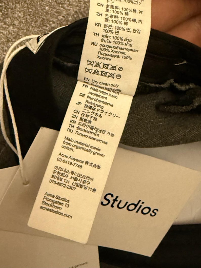 May include: A white fabric care label attached to a black garment, featuring multi-language text including 'Dry clean only' and 'Main material made from organically grown cotton.' A separate white paper tag displays the brand name 'Studios' in black.