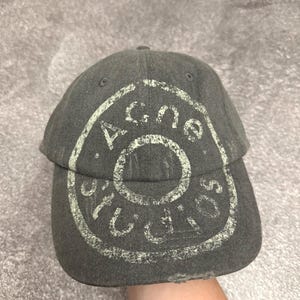 May include: A charcoal gray baseball cap featuring a distressed, light-colored circular graphic print on the front panel. The text "Acne Studios" is printed in a circular pattern around the center. The brim shows signs of wear with frayed edges.