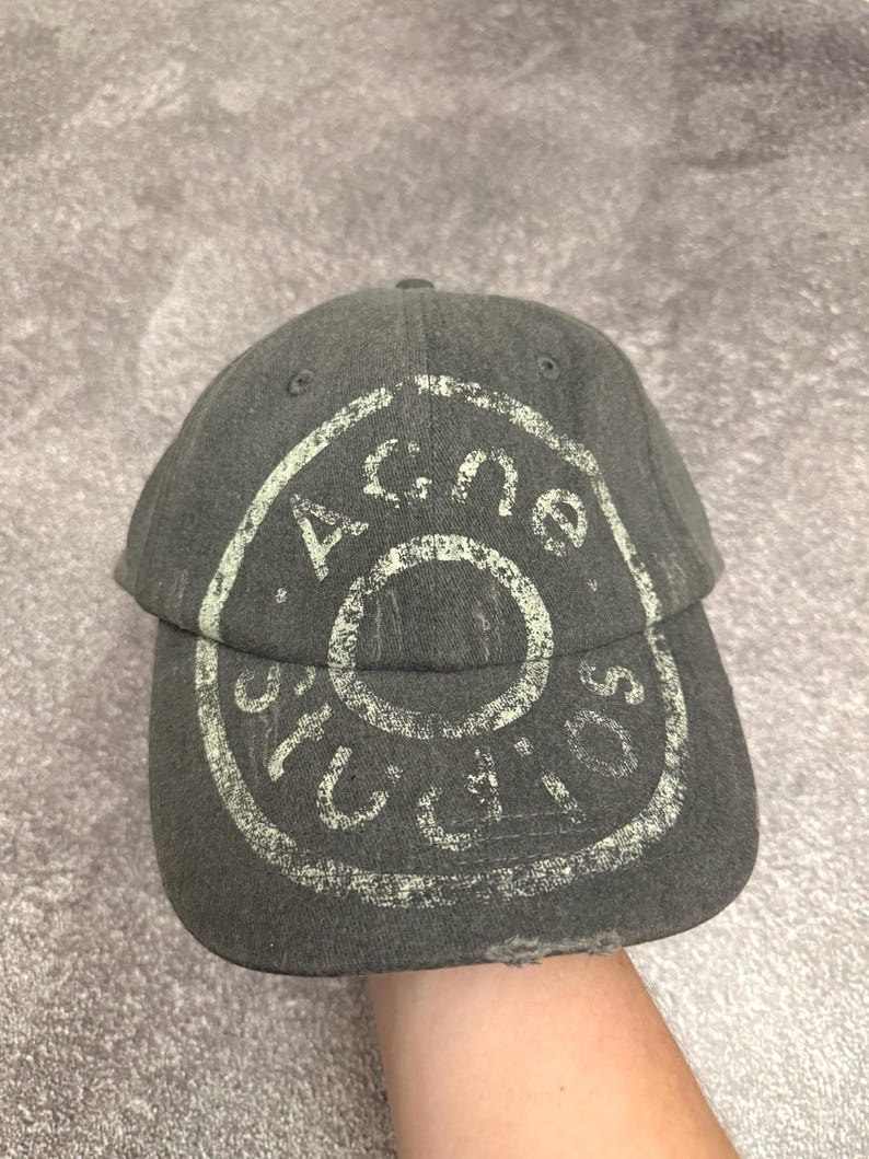 May include: A charcoal gray baseball cap featuring a distressed, light-colored circular graphic print on the front panel. The text "Acne Studios" is printed in a circular pattern around the center. The brim shows signs of wear with frayed edges.