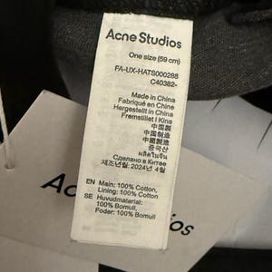 May include: A white fabric care label attached to a black garment. The label features the text Acne Studios, One size (59 cm), FA-UX-HATS000288, C40382-, Made in China, and specifies the composition as 100% Cotton for both the main and lining materials.