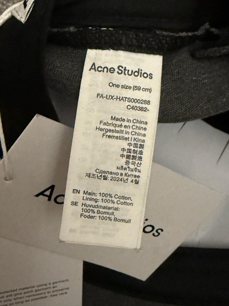 May include: A white fabric care label attached to a black garment. The label features the text Acne Studios, One size (59 cm), FA-UX-HATS000288, C40382-, Made in China, and specifies the composition as 100% Cotton for both the main and lining materials.