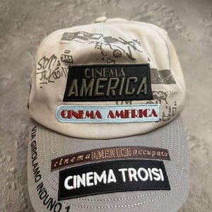 Może przedstawiać: Dwukolorowa czapka bejsbolowa z szarym daszkiem i beżową koroną. Czapka posiada napis "CINEMA AMERICA" na czarnej naszywce oraz inne teksty i grafiki. Daszek ma napis "CINEMA TROISI".