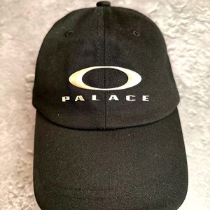 Puede incluir: Una gorra de béisbol negra con un logotipo ovalado de color dorado metálico centrado en la parte frontal. Debajo del óvalo, la palabra PALACE está impresa en una fuente dorada metálica a juego. La gorra tiene un diseño estructurado de seis paneles.