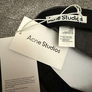 May include: A dark grey baseball cap featuring a white interior brand label reading Acne Studios, a white hang tag with the text Acne Studios, and a care instruction tag detailing 100% cotton composition and garment care information.