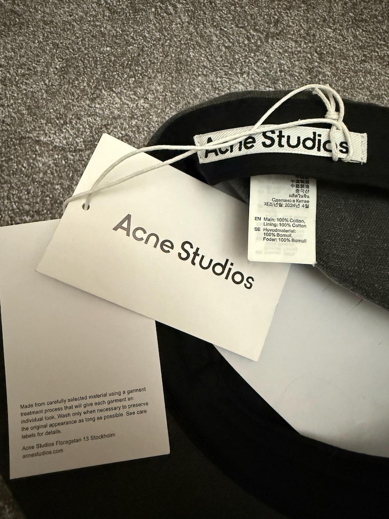 May include: A dark grey baseball cap featuring a white interior brand label reading Acne Studios, a white hang tag with the text Acne Studios, and a care instruction tag detailing 100% cotton composition and garment care information.