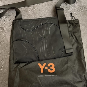 Peut inclure: Sac bandouli&egrave;re noir avec une longue sangle, motif ondul&eacute; et logo Y-3. Comprend un sac de rangement noir avec le logo Y-3 et le texte "adidas YOHJI YAMAMOTO" en orange.