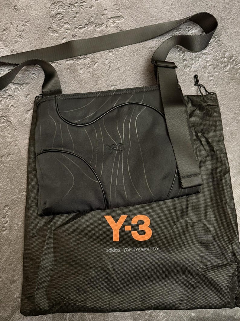 Peut inclure: Sac bandouli&egrave;re noir avec une longue sangle, motif ondul&eacute; et logo Y-3. Comprend un sac de rangement noir avec le logo Y-3 et le texte "adidas YOHJI YAMAMOTO" en orange.