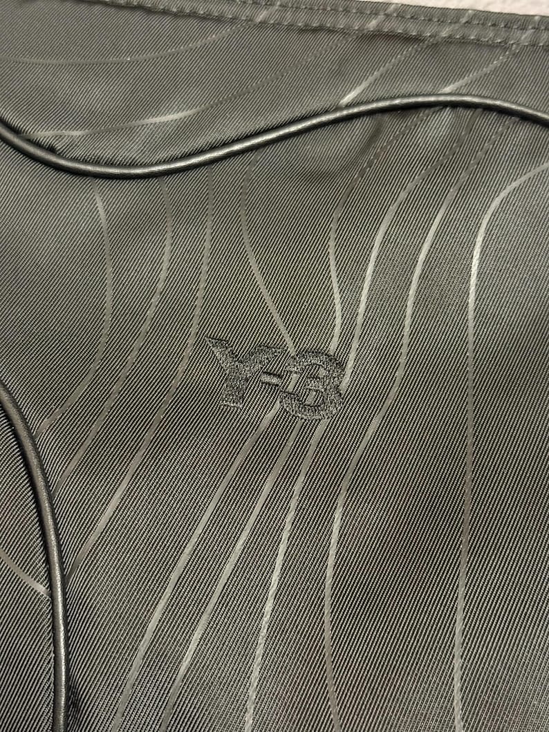 Peut inclure: Gros plan sur un tissu vert fonc&eacute; avec un motif &agrave; rayures verticales. Le logo Y-3 est brod&eacute; en bas au centre. Une bordure noire et arrondie est visible en haut et &agrave; gauche de l'image.