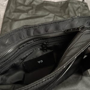 Peut inclure: Sac &agrave; bandouli&egrave;re noir avec un compartiment principal zipp&eacute; et une petite poche int&eacute;rieure. Le sac porte le logo "Y-3" sur une &eacute;tiquette noire. Un sac en tissu noir assorti est visible en arri&egrave;re-plan.