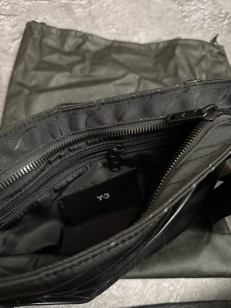 Peut inclure: Sac &agrave; bandouli&egrave;re noir avec un compartiment principal zipp&eacute; et une petite poche int&eacute;rieure. Le sac porte le logo "Y-3" sur une &eacute;tiquette noire. Un sac en tissu noir assorti est visible en arri&egrave;re-plan.