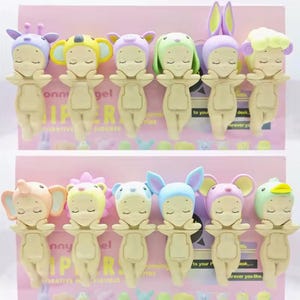 May include: A collection of small, whimsical figurines, each dressed in a different animal-themed hat. The figurines are in various pastel colors, including pink, yellow, and blue. The text "Sunny Angel" is visible on the packaging.