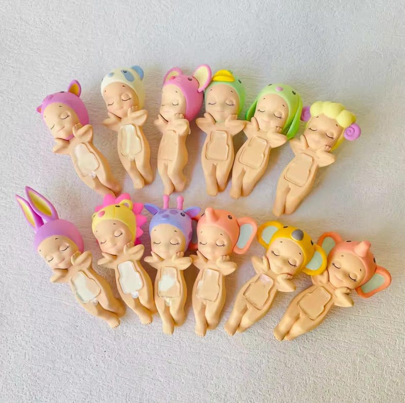 May include: A collection of small, sleeping baby figurines with various animal-themed hats. The figurines are in a lying down position, with closed eyes and arms outstretched. The hats include bunny ears, bear ears, and other animal designs. The color palette is soft, with pastel shades.