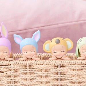 May include: Four small figurines in a woven basket, each wearing a different animal-themed hat: a bunny, a koala, and two others. The figurines are in pastel colors, with closed eyes, resting on their arms.