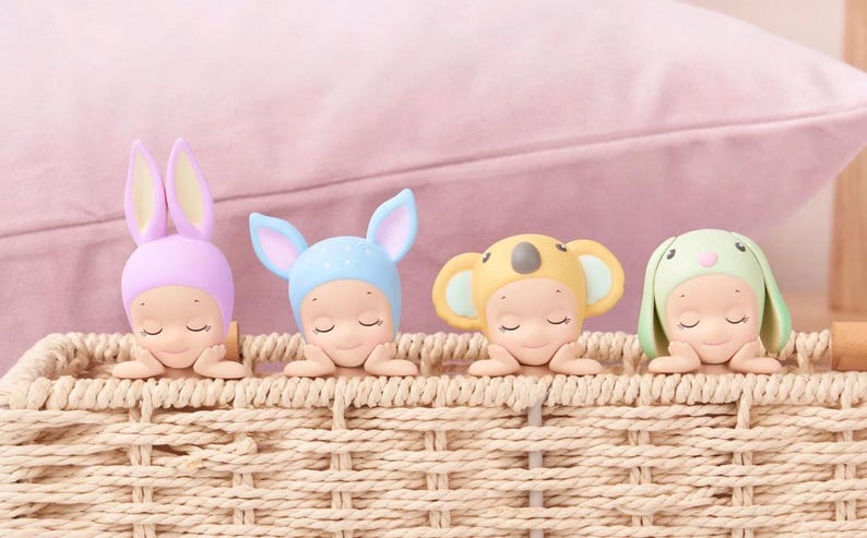 May include: Four small figurines in a woven basket, each wearing a different animal-themed hat: a bunny, a koala, and two others. The figurines are in pastel colors, with closed eyes, resting on their arms.