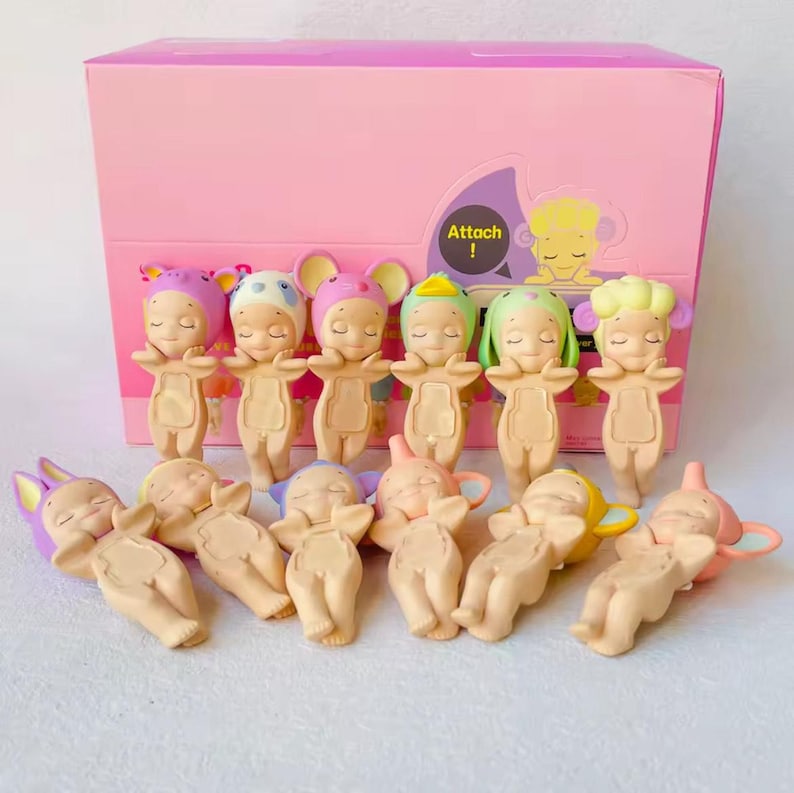 May include: A collection of small, sleeping baby figurines with various animal-themed hats, arranged in front of a pink box. The figurines are beige with closed eyes and outstretched arms. The box has the word "Attach" on it.