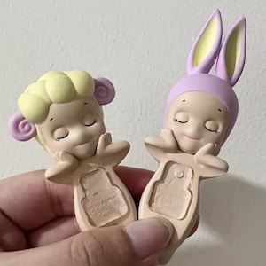 May include: Two small, pastel-colored figurines. One is a sheep, the other a bunny. Both have closed eyes and are resting their chins on their hands. The sheep has yellow hair and purple accents. The bunny has a purple hood and yellow ears.